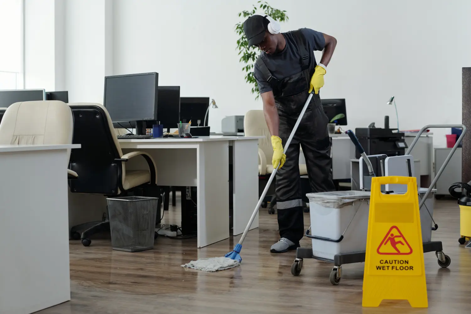 Zeitgenössischer junger schwarzer Mann in Arbeitskleidung reinigt vorsichtig den Boden im Großraumbüro vor einem gelben Plastikschild